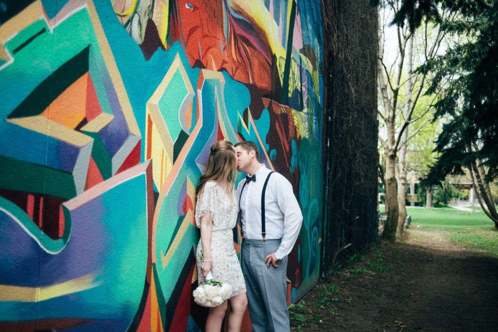 bride is leaning up against a graffiti wall and the groom is leaning in to kiss her