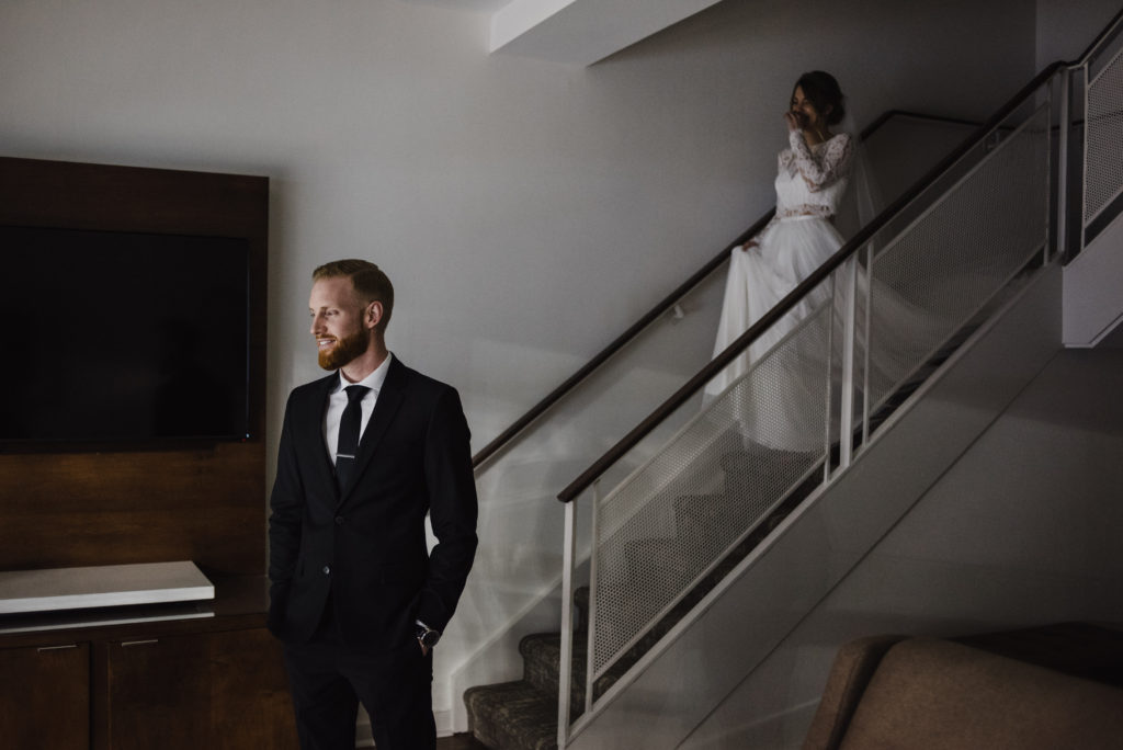 groom is standing at the bottom of the stairs looking away and the bride is walking down the stairs towards him to do a first look