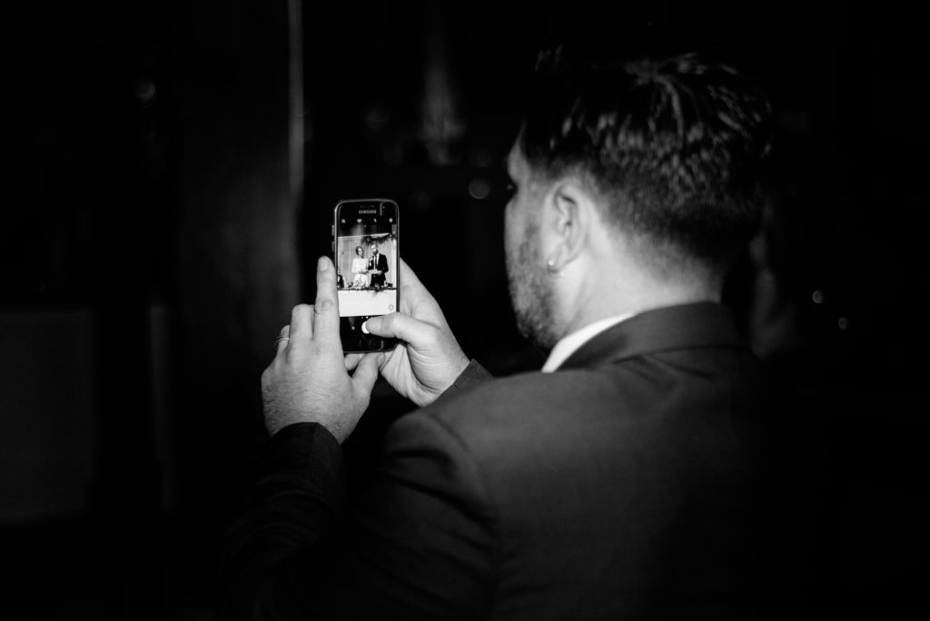 black and white photo of a guests iPhone while he is taking a photo of the bride and grooms speech