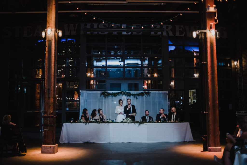 photo of entire head table at steam whistle brewery while bride and groom are giving their speech