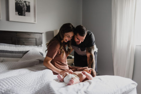 new mother and father sitting on the edge of a bed looking down at their new baby girl who is laying on the bed