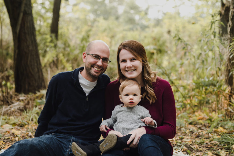 Family picture of a father, mother, and infant daughter sitting in the fall leaves for a picture